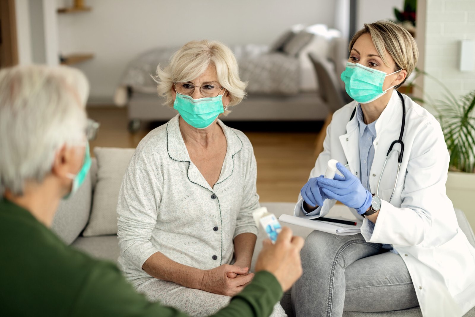 Female doctor talking to senior couple while vising them at home during coronavirus epidemic. They are wearing protective face masks.