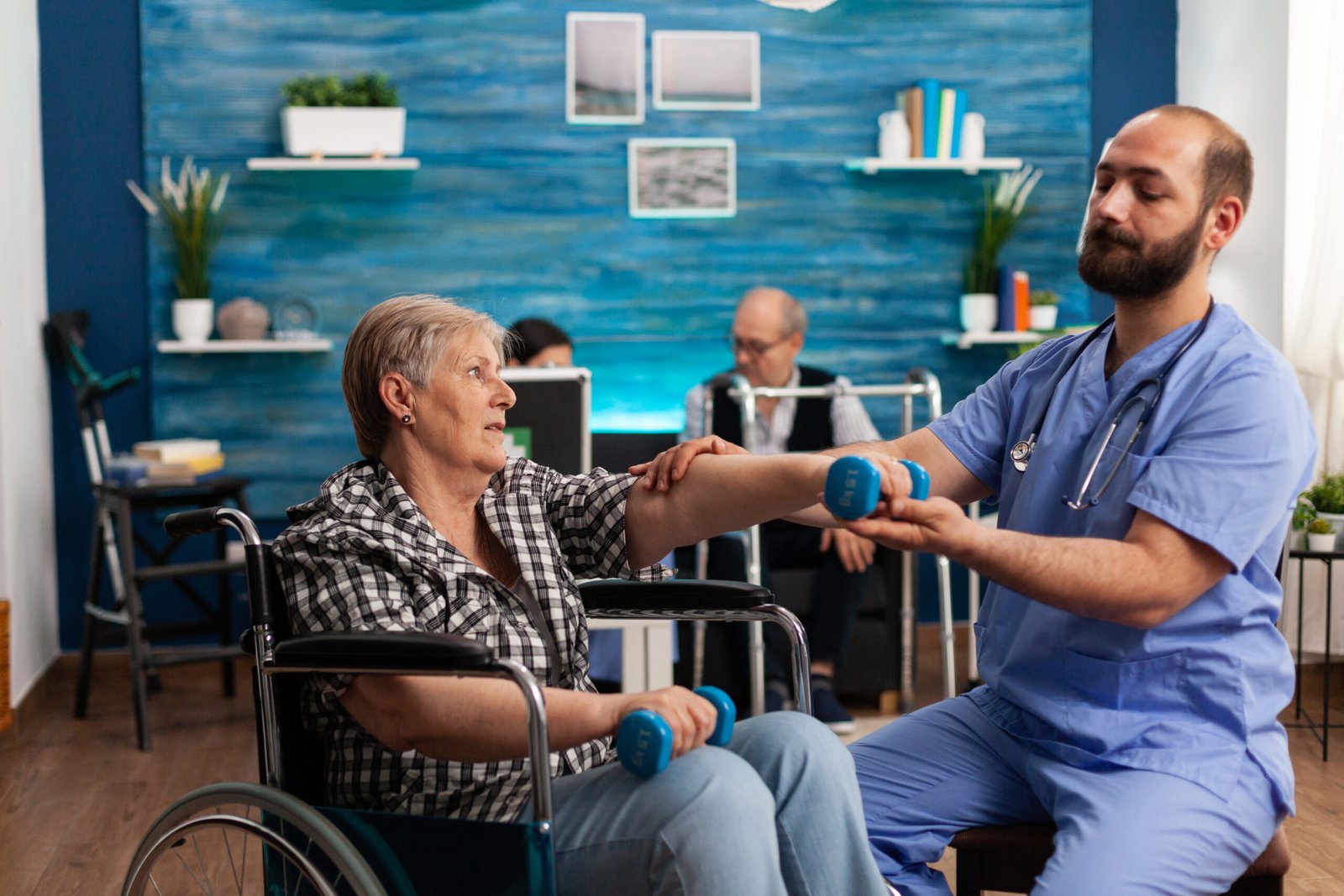 Male nurse helping senior retired disabled woman in wheelchair to rehabilitate using dumbbels during recovery session in nursing home. Elderly woman exercising physiotherapy with social worker